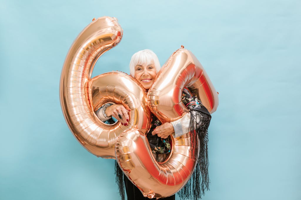 Happy senior woman poses with 64 balloons on a light blue background, exuding joy and celebration.